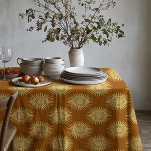 Puede incluir: Una mesa de comedor con un mantel mostaza con un diseño de sol. La mesa está puesta con platos, cuencos, vasos, un jarrón con flores y fruta. Se ve parcialmente una silla de madera.