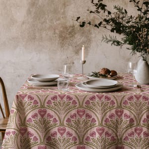 May include: A dining table set for a meal, featuring a pink tablecloth with a repeating heart and leaf pattern. The table is set with plates, glasses, and a vase of flowers. A lit candle adds ambiance.