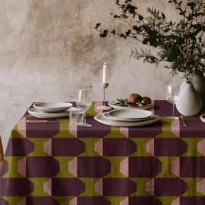 May include: A dining table set with a patterned tablecloth in burgundy, olive green, and blush pink. The table is set with white plates, glasses, and a candle. A vase of flowers and a wooden chair are also visible. The tablecloth has a geometric design.