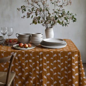 Puede incluir: Una mesa de comedor preparada para una comida, con un mantel color mostaza con un estampado floral blanco. La mesa está puesta con platos, cuencos, vasos y un jarrón con flores. Una silla de madera está en primer plano.