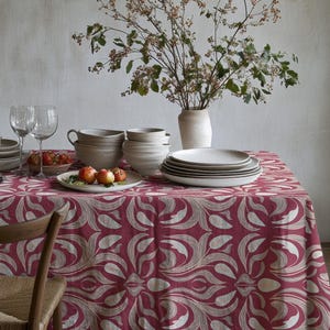 Peut inclure: Une table de salle à manger dressée pour un repas, avec une nappe bordeaux à motifs floraux blancs. La table est garnie d'assiettes, de bols, de verres et d'un vase de branches. Une chaise en bois est partiellement visible.