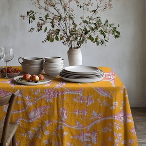 Op de afbeelding: Een eettafel gedekt met een gele tafelkleed met een roze patroon. De tafel is gedekt met borden, kommen, glazen en een vaas met bloemen. Een houten stoel is gedeeltelijk zichtbaar.