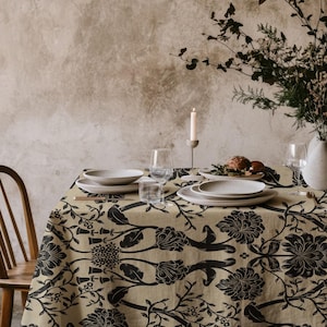 Könnte beinhalten: Ein Esstisch für eine Mahlzeit, mit einer beigefarbenen Tischdecke mit schwarzem Blumenmuster. Der Tisch ist mit weißen Tellern, Gläsern und einer Blumenvase gedeckt. Eine brennende Kerze sorgt für Ambiente.
