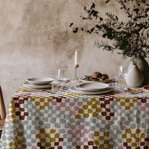Op de afbeelding: Een eettafel gedekt voor een maaltijd, met een patroon tafelkleed in rood, geel, blauw en roze. De tafel is gedekt met borden, glazen en een vaas met bloemen. Een brandende kaars voegt sfeer toe.