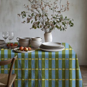 May include: A dining table set for a meal, featuring a blue and green striped tablecloth. The table is set with plates, bowls, glasses, and a vase of flowers. A wooden chair is in the foreground. The scene is lit with natural light.