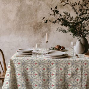 Può includere: Un tavolo da pranzo apparecchiato per un pasto, con una tovaglia color crema con un motivo floreale ripetuto in rosso e blu. Il tavolo è apparecchiato con piatti, bicchieri e un vaso di fiori. Una candela accesa contribuisce all'atmosfera.