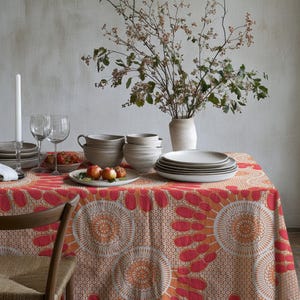 Puede incluir: Una mesa de comedor decorada con un mantel estampado en rojo y naranja. La mesa está puesta con platos, cuencos, vasos, una vela encendida y un jarrón con ramas. También se ve una silla de madera.