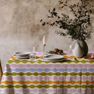 Op de afbeelding: Een eettafel gedekt voor een maaltijd, met een kleurrijk tafelkleed met een golvend patroon in roze, groen, oranje en paars. De tafel is gedekt met borden, glazen en een vaas met bloemen. Een houten stoel is zichtbaar.