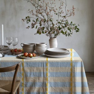 Op de afbeelding: Een eettafel gedekt voor een maaltijd, met een blauw en geel gestreept tafelkleed. De tafel is gedekt met borden, kommen, glazen en een vaas met bloemen. Een houten stoel staat op de voorgrond. Een brandende kaars staat links.