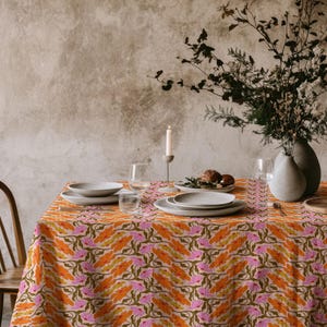 Puede incluir: Una mesa de comedor con un mantel estampado en naranja y rosa. Se colocan platos, vasos y un jarrón con flores. Una silla de madera es visible en el lado izquierdo. La escena está lista para una comida.
