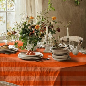 May include: A dining table set with an orange and white striped tablecloth. White plates, silverware, and wine glasses are arranged on the table. A floral arrangement with orange and yellow flowers is the centerpiece, with candles.