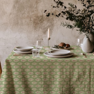Puede incluir: Una mesa de comedor preparada para una comida, con un mantel verde con un patrón de hojas repetido en rojo y crema. La mesa está puesta con platos blancos, vasos y un jarrón de flores. Una silla de madera está a la izquierda.