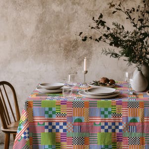 Puede incluir: Un mantel colorido con un patrón geométrico de cuadrados, rayas y formas en varios colores, incluyendo rosa, naranja, verde y azul. La mesa está puesta con platos, vasos y un jarrón con flores, creando una escena vibrante.