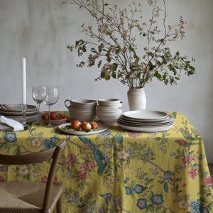 Könnte beinhalten: Ein gedeckter Esstisch mit gelber Blumendecke, Keramikplatten, Schalen und Gläsern. Eine Vase enthält Zweige mit kleinen Blüten. Äpfel und Erdbeeren liegen auf Tellern.