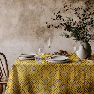 Può includere: Una tovaglia con motivo giallo e blu con un design geometrico. La tovaglia è apparecchiata con piatti, ciotole e bicchieri. Un vaso con rami secchi si trova sul tavolo.