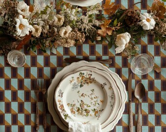 Peut inclure: Un couvert de table avec une nappe à motifs géométriques bleu sarcelle, marron et crème. L'ensemble comprend des assiettes en céramique blanche à motifs floraux, des fourchettes, une cuillère et un couteau en argent, des verres et des fleurs séchées.