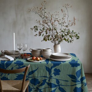 May include: A sage green tablecloth featuring a bold dark blue floral pattern, set with a stack of ceramic plates, bowls, a vase with dried branches, two wine glasses, a white taper candle, and a small bowl containing several red apples.