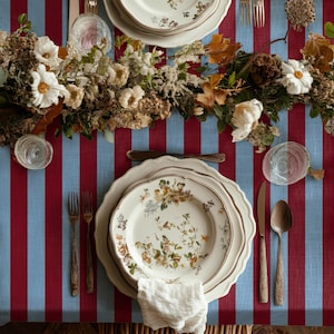 May include: A table setting with floral patterned plates, silverware, and a white linen napkin. The table is covered with a red and light blue striped tablecloth. A floral arrangement with white flowers and dried leaves decorates the table.