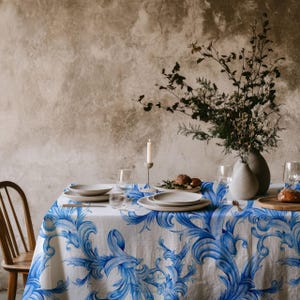 Puede incluir: Un mantel blanco con un patrón floral azul y blanco, puesto con platos blancos, vasos y un jarrón con flores secas. El mantel está sobre una mesa frente a una pared texturizada.