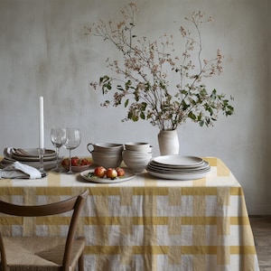 May include: A dining table covered with a yellow and white checkered tablecloth, featuring a stack of ceramic plates, bowls, two wine glasses, a white taper candle, a small bowl of red fruit, and a vase with tall leafy branches.