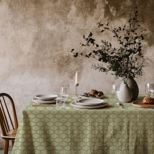May include: A sage green tablecloth featuring a repeating pattern of small, stylised leaf motifs in muted pink and cream. The table is set with white ceramic plates, clear glassware, a tall taper candle, and a vase filled with dried branches.