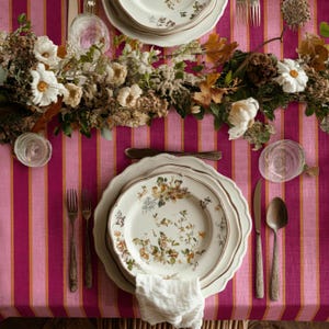 May include: A table setting with floral patterned plates, silverware, and a white napkin. The table is covered with a pink and orange striped tablecloth. A floral arrangement with white flowers and greenery decorates the table.
