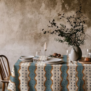 May include: A rectangular tablecloth featuring a repeating vertical pattern of blue, cream, and mustard-yellow scalloped stripes with small dark blue star motifs. The fabric is draped over a table set with white ceramic plates, glassware, and a vase.