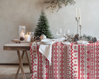 May include: A rectangular tablecloth featuring a repeating vertical pattern of red snowflakes, hearts, candy canes, and pine trees on a cream-colored background. The fabric drapes over a wooden table set with white plates, clear glasses, and candles.