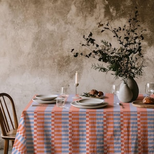 May include: A rectangular tablecloth featuring a repeating geometric pattern of vertical stripes in shades of orange, light blue, and soft pink. The fabric drapes over a dining table set with white ceramic plates, clear glassware, and a candle.