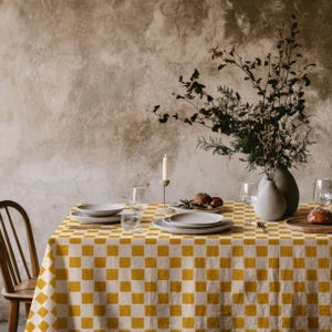 Puede incluir: Una mesa de comedor con un mantel a cuadros amarillos y blancos. La mesa está puesta con platos, vasos y un jarrón con vegetación. También se ven una vela encendida y una silla de madera.