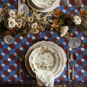 May include: A dining table set with floral plates, cutlery, and a white napkin. The table is covered with a blue and burgundy checked tablecloth with white starbursts. A floral centerpiece adds a natural touch.