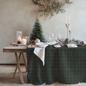 May include: A rectangular tablecloth featuring a dark green and navy blue plaid pattern draped over a wooden table. The table is set with white plates, clear wine glasses, white napkins, white pillar candles in glass holders, and pinecones.