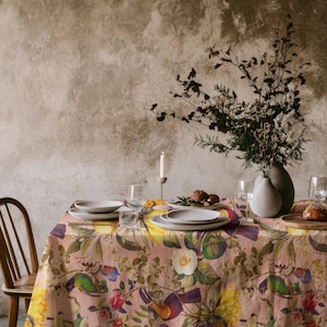 May include: A rectangular tablecloth featuring a botanical and avian print with yellow sunflowers, green leaves, and colourful birds on a dusty pink background. The table is set with white ceramic plates, clear glasses, and a tall white candle.