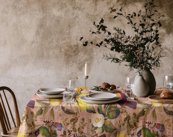 May include: A rectangular tablecloth featuring a botanical and avian print with yellow sunflowers, green leaves, and colorful birds on a dusty pink background. The table is set with white ceramic plates, clear glasses, and a tall white candle.