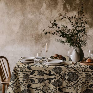 May include: A dining table covered with a beige tablecloth featuring a black floral pattern. The table is set with white ceramic plates, clear glass stemware, a tall candle in a holder, and a large vase filled with dried branches and greenery.