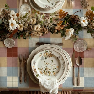 May include: A plaid tablecloth in muted tones of blue, yellow, and red features a place setting with a floral-patterned dinner plate, a white napkin, two forks, a knife, a spoon, and two clear glasses, accented by a centrepiece of dried flowers.