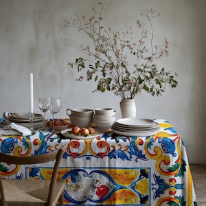 May include: A white tablecloth with a colourful floral pattern features a set of white ceramic bowls and plates, two wine glasses, and a white candle. A bouquet of dried branches in a white vase sits on the table.
