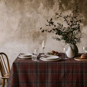 May include: A dark red and black plaid tablecloth covers a dining table set with white ceramic plates, clear glass stemware, a single white taper candle in a holder, and a large ceramic vase containing dried branches and foliage.