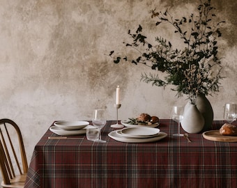 May include: A dark red and black plaid tablecloth covers a dining table set with white ceramic plates, clear glass stemware, a single white taper candle in a holder, and a large ceramic vase containing dried branches and foliage.