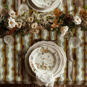 May include: A table setting featuring a patterned tablecloth with wavy vertical stripes in olive green, brown, and cream. The setting includes white ceramic plates with floral motifs, clear glass tumblers, metal forks, and spoons.