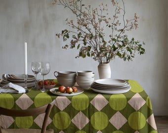 Peut inclure: Une nappe rectangulaire ornée d'un motif géométrique répétitif composé de cercles vert olive et de losanges rose pâle. La table est dressée avec une pile d'assiettes et de bols en céramique blanche, une bougie blanche, deux verres et des pommes.
