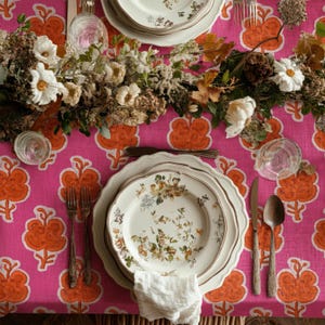 May include: A bright pink tablecloth with an orange floral pattern features a place setting with white ceramic plates, a white napkin, silver forks, a silver knife, and a silver spoon, decorated with white flowers and dried foliage.