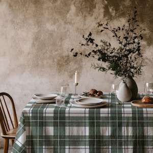 May include: A rectangular tablecloth featuring a green, white, and dark blue plaid pattern. The table is set with white ceramic plates, clear glass stemware, a single white taper candle in a holder, and a vase containing dried branches.