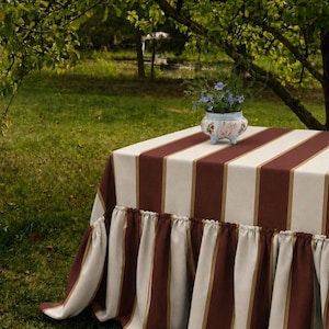 May include: A rectangular tablecloth featuring vertical stripes in shades of deep maroon, cream, and thin gold lines. The tablecloth includes a gathered, ruffled skirt along the bottom edge and supports a small, light-colored ceramic bowl with floral details.