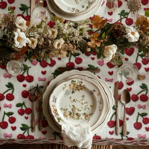 May include: A beautifully set table with floral patterned plates, silverware, and a white napkin. The tablecloth is white with a cherry and bow pattern. A floral centerpiece adds a touch of elegance to the dining setting.