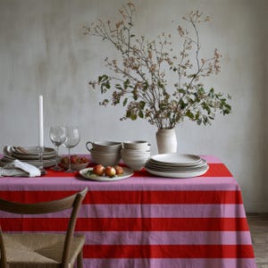 May include: A rectangular tablecloth featuring horizontal stripes in alternating shades of bright red and light pink. The table is set with stacks of neutral-toned ceramic plates and bowls, two clear glass wine glasses, a white candle, and a small bowl of fruit.