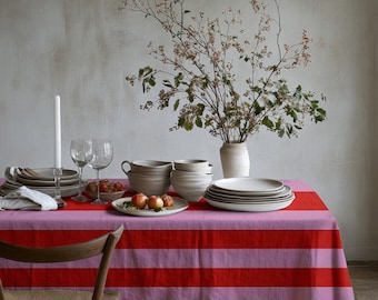 May include: A rectangular tablecloth featuring horizontal stripes in alternating shades of bright red and light pink. The table is set with stacks of neutral-toned ceramic plates and bowls, two clear glass wine glasses, a white candle, and a small bowl of fruit.