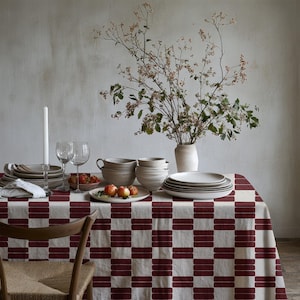 Peut inclure: Une table à manger recouverte d'une nappe à carreaux crème et rouge foncé. La table est dressée avec des piles de bols et d'assiettes en céramique crème, une bougie blanche, deux verres à vin transparents, un bol de fruits rouges et un vase.