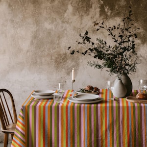 May include: A rectangular tablecloth featuring vertical stripes in shades of orange, pink, light blue, olive green, and brown. The table is set with white ceramic plates, clear glass stemware, a single white taper candle, and a vase with dried branches.