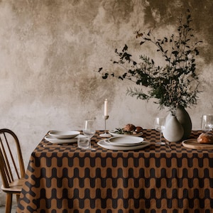 May include: A rectangular tablecloth featuring a repeating dark brown and tan wavy geometric pattern. The table is set with white ceramic plates, clear glass stemware, a single white taper candle in a holder, and a vase filled with dried branches.